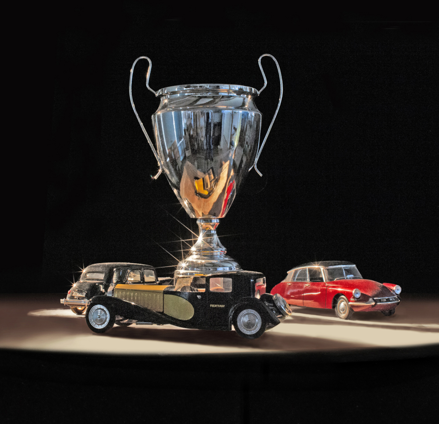 Trois maquettes de voitures anciennes dont une Citroën DS rouge, entourant une coupe trophée chromée brillante